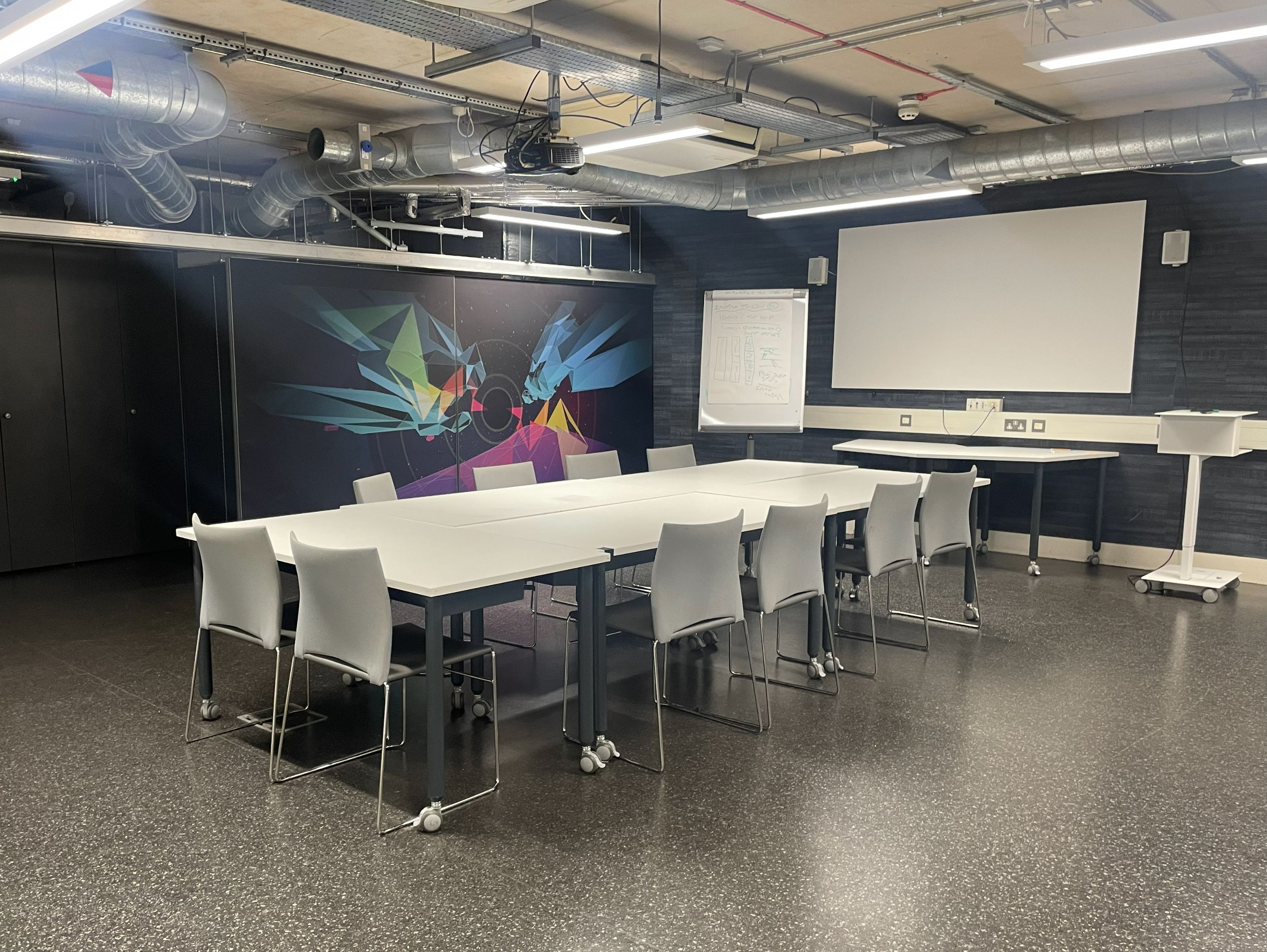 A room with a large, configurable table with chairs, plus a projector and a flip board