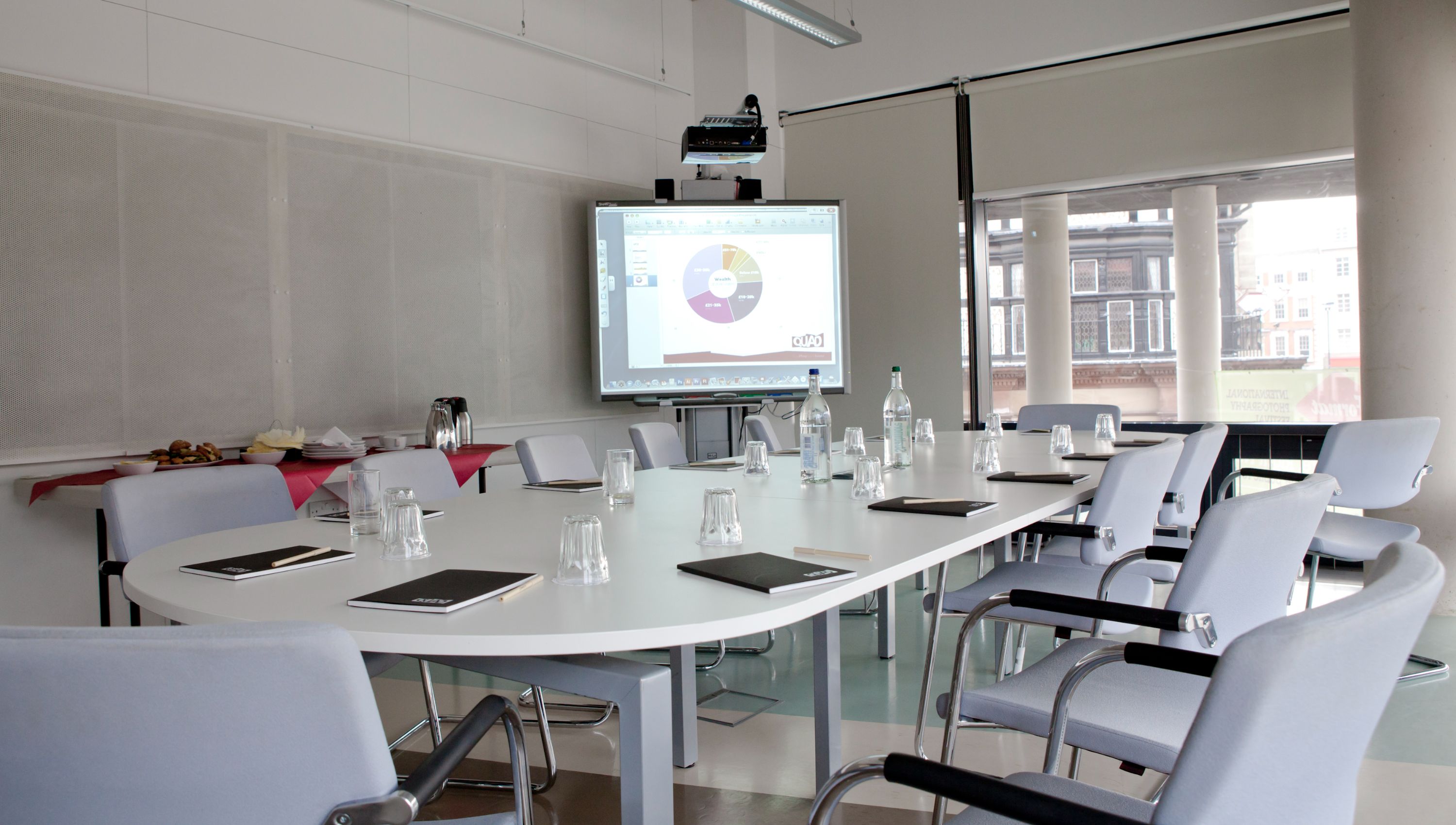 Image Credit: Graham Lucas Commons A meeting room with a large table set with water glasses and notebooks, angled towards a projection screen